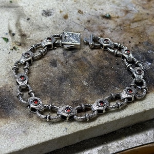 May include: A silver chain bracelet with a detailed engraved design and red gemstone accents.