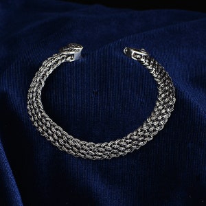 May include: Silver braided bangle bracelet with a decorative clasp. The bracelet is on a dark blue fabric background.
