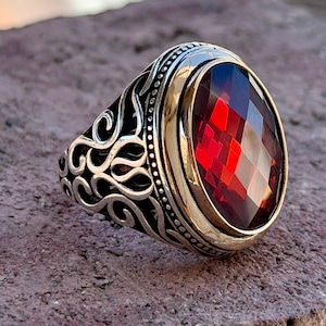 Op de afbeelding: Een zilveren ring met een ovale, gefacetteerde dieprode steen in een goudkleurige zetting. De band is voorzien van ingewikkelde, donker geoxideerde krulpatronen die langs de zijkanten lopen, wat zorgt voor een contrastrijke, textuurrijke uitstraling.