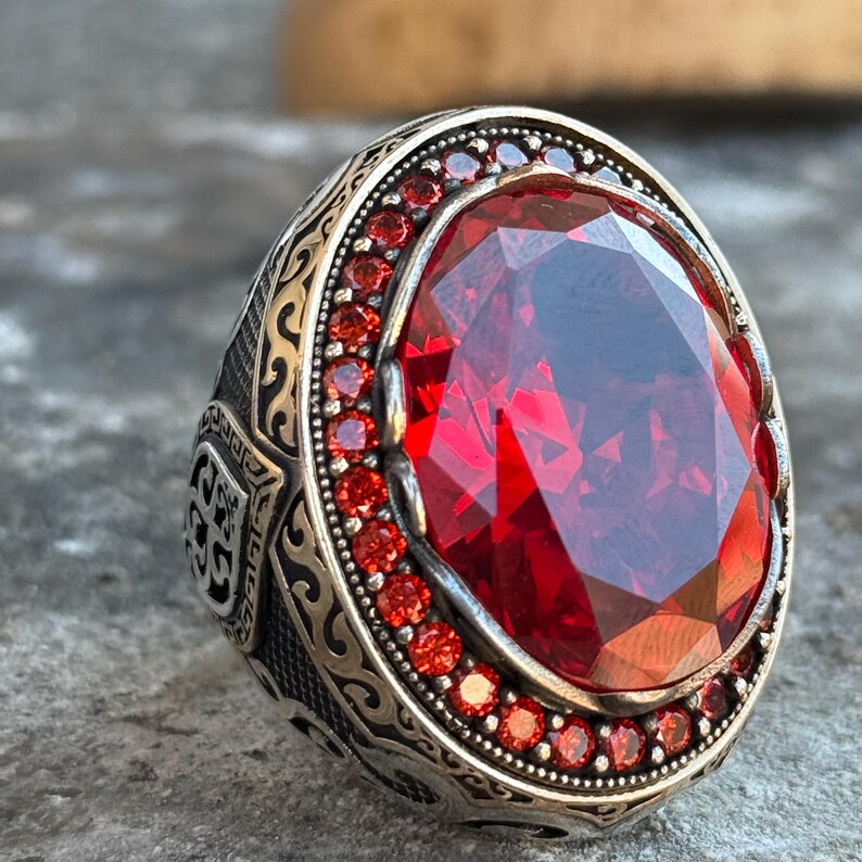 Puede incluir: Anillo de plata con una piedra preciosa roja oscura ovalada y facetada como pieza central. El anillo est&aacute; rodeado de piedras rojas m&aacute;s peque&ntilde;as y presenta intrincados detalles grabados en la banda.