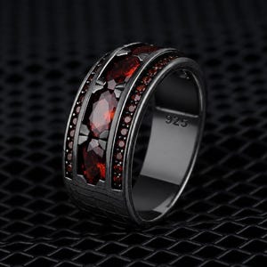 May include: A black metal ring featuring a central row of three marquise-cut deep red gemstones flanked by two rows of small red stones. The band has a textured grid pattern on the sides and is stamped with the number 925 on the inner surface.