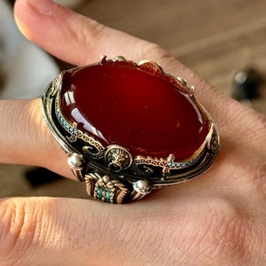 May include: A silver ring with an ornate design featuring a red carnelian stone. The ring has a lion head detail and turquoise accents.
