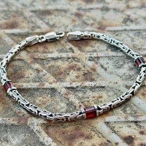 Peut inclure: Bracelet argenté avec un design de chaîne détaillé et trois accents de pierres précieuses rouges rectangulaires. Le bracelet est présenté sur une surface métallique rouillée et texturée, soulignant son savoir-faire et son contraste de couleurs.
