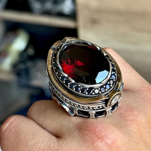 May include: A silver ring with a large, oval, deep red gemstone as the centerpiece. The ring has a detailed design with black accents around the gemstone and a patterned band. The ring is a statement piece.