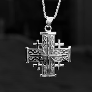 May include: A silver Jerusalem cross pendant with an intricate design, hanging from a matching chain. The cross has smaller crosses at each arm and decorative scrollwork. The background is a dark, blurred gradient.