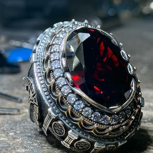 May include: A detailed silver ring featuring a large, faceted, deep red gemstone as the centerpiece. The ring is adorned with smaller, clear gemstones and intricate metalwork around the main stone, showcasing a vintage or antique style.