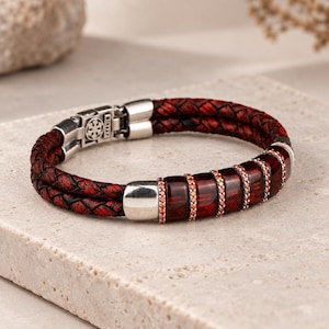 May include: A double-strand braided dark red leather bracelet featuring a silver-toned metal clasp and a series of five rectangular dark red resin segments separated by thin bands of small, sparkling orange-red stones.
