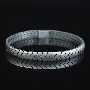 Puede incluir: Un brazalete de metal en tono plateado que presenta un patrón repetitivo de chevrón o flecha a lo largo de toda su circunferencia. El brazalete tiene un acabado mate cepillado y un ancho constante, creando una apariencia geométrica estructurada.