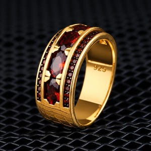 May include: A gold-toned ring featuring a central row of three oval-cut deep red gemstones flanked by two rows of smaller round red stones. The band has a textured grid pattern on the lower section and the engraving 925 on the inner surface.