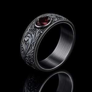 Könnte beinhalten: Ein dunkelgrauer Metallring mit einer detaillierten floralen Gravur auf dem äußeren Band. Der Ring ist mit einem einzelnen, runden, tiefroten Edelstein in einer glatten Fassung besetzt. Die Kanten des Bandes haben eine feine Perlenstruktur.