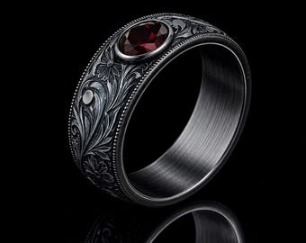 May include: A dark grey metal ring featuring an intricate floral engraving across the outer band. The ring is accented with a single, round, deep red gemstone set in a smooth bezel. The edges of the band are finished with a fine beaded texture.