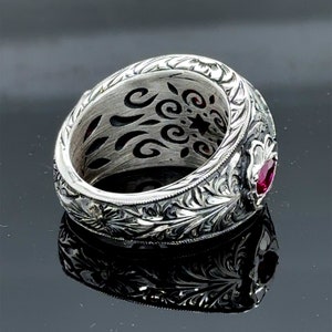 Peut inclure: Une bague en argent avec des gravures florales complexes et une seule pierre pr&eacute;cieuse rouge sertie dans la bande.