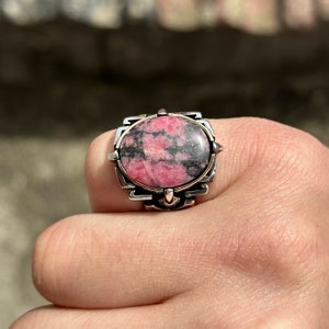 May include: A silver ring with a pink and black oval-shaped gemstone set in a detailed silver setting.