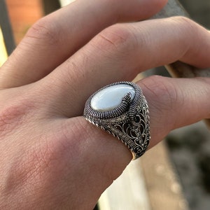 Puede incluir: Un anillo de plata con una piedra ovalada blanca y detalles filigranados intrincados. El anillo se lleva en el dedo de una persona.