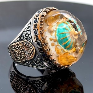 May include: A silver ring with a large, clear, faceted gemstone. The gemstone contains a gold and turquoise design. The ring band is decorated with intricate details and black stones.