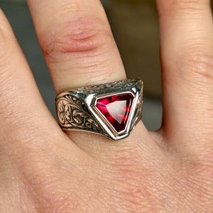 Puede incluir: Un anillo de plata con un diseño intrincado y una piedra preciosa triangular roja.