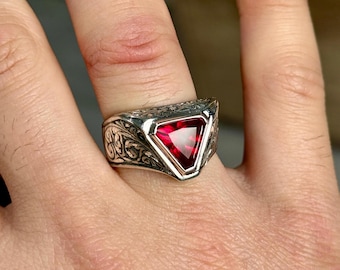 May include: A silver ring with an intricate design and a red triangular gemstone.