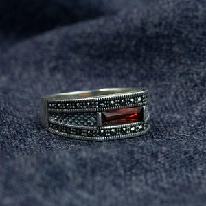 May include: A silver ring featuring a rectangular, deep red gemstone. The band is adorned with small, dark embellishments, giving it a vintage appearance. The ring is set against a dark, textured backdrop, suggesting a focus on the jewellery.