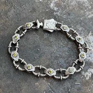 May include: Silver bracelet featuring a series of ornate, diamond-shaped links, each housing a small, round, green gemstone. The bracelet has a decorative clasp with engraved floral patterns. The links are connected by smaller, oval-shaped links.