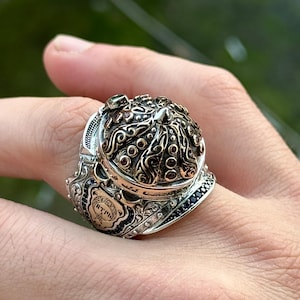 Pode incluir: Um anel de prata com um design detalhado e ornamentado. O anel apresenta uma parte superior em forma de cúpula com entalhes intrincados e uma pequena ponta pontiaguda. A cúpula é cercada por uma banda de pedras preciosas pretas. O anel está colocado em um dedo.