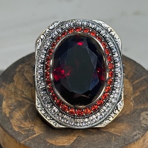 May include: A silver ring featuring a large, oval, dark red gemstone. The gemstone is encircled by a row of small red stones and a row of clear stones. The ring has an intricate, detailed design, with an ornate setting.