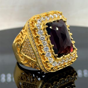 Pode incluir: Um anel dourado com uma grande pedra retangular vermelho escuro. O anel é adornado com pedras preciosas menores e transparentes e detalhes intrincados em filigrana. A moldura quadrada tem uma borda decorativa.