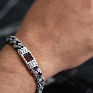 May include: Silver bracelet with a square, red gemstone centerpiece. The bracelet features a patterned chain design with a decorative clasp. The jewelry is displayed on a person's wrist, showcasing the details of the accessory.