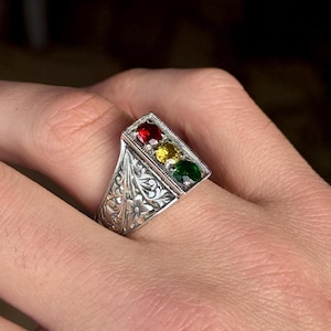 Puede incluir: Anillo de plata con una parte superior rectangular con tres piedras preciosas: roja, amarilla y verde. El anillo tiene un intrincado diseño floral en la banda. Las piedras están engastadas en un marco rectangular.