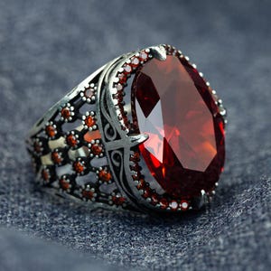May include: A silver ring with a large, faceted, oval-shaped red gemstone. The ring has intricate filigree detailing and smaller red gemstones around the main stone, creating a striking contrast.