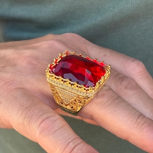Könnte beinhalten: Ein goldfarbener Ring mit einem großen, rechteckigen, tiefroten Edelstein. Der Ring hat ein verziertes Design mit kleineren, klaren Steinen und detaillierter Filigranarbeit. Die Fassung ist goldfarben und erzeugt ein luxuriöses Aussehen.