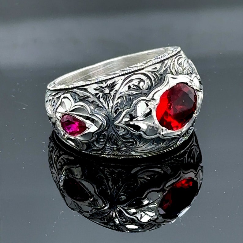Peut inclure: Une bague en argent avec des motifs floraux complexes et deux pierres pr&eacute;cieuses rouges. La bague est large et a un profil haut.
