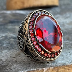 May include: A silver ring featuring a large, oval-cut, deep red gemstone. The ring is encircled by smaller red stones and intricate silver filigree. The band displays engraved patterns, giving it a vintage or antique appearance.