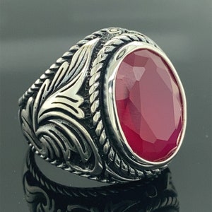 Può includere: Un anello d'argento con un designo ornato che presenta una grande pietra preziosa rossa sfaccettata.