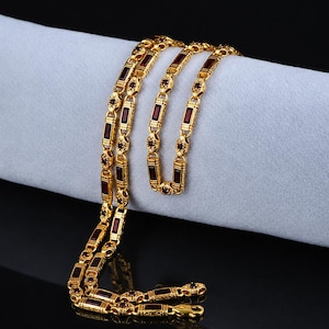 Puede incluir: Un collar dorado con piedras rojas rectangulares. La cadena se muestra sobre una superficie gris claro, con un fondo negro. Los eslabones del collar están detallados con patrones ornamentados.