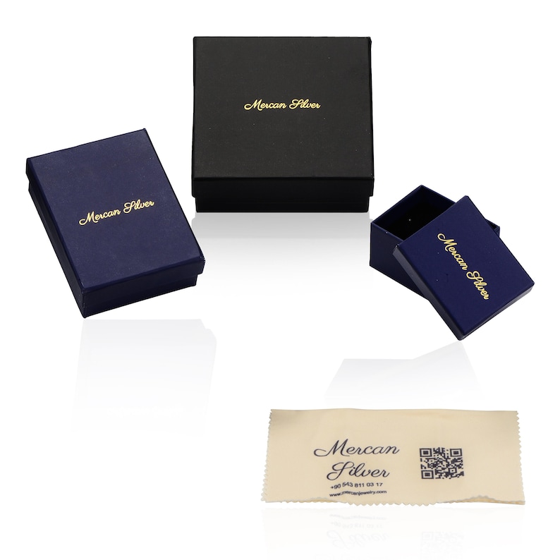 Puede incluir: Varias cajas de joyer&iacute;a en azul marino y negro, con la inscripci&oacute;n dorada "Mercan Silver". Una de las cajas est&aacute; abierta, mostrando su interior. Tambi&eacute;n se incluye un pa&ntilde;o de limpieza beige con el mismo logotipo y un c&oacute;digo QR.