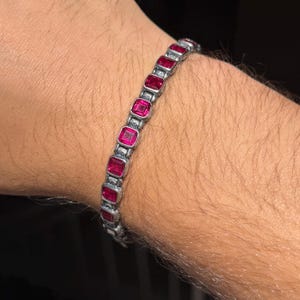 Puede incluir: Una pulsera de eslabones en tono plateado con una serie de piedras preciosas de color rojo rosado, de corte cuadrado y engastadas en marcos metálicos individuales. La pulsera se compone de eslabones rectangulares que conectan cada engaste.