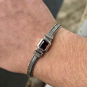 May include: A silver bracelet with a rectangular, deep red gemstone at the center. The bracelet has a braided design on the band and decorative square accents on either side of the gemstone. The bracelet is worn on a wrist.