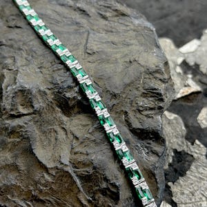 May include: A silver bracelet with alternating emerald green and clear rectangular stones. The bracelet is displayed on a dark, textured surface, highlighting the sparkle of the gemstones. The bracelet is a piece of jewelry.