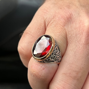 May include: A silver ring with an ornate design and a large, oval-shaped red gemstone.