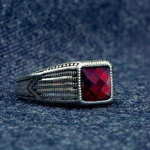 May include: A silver ring with a large, square, faceted, red gemstone. The ring has a detailed, textured design on the band and around the gemstone setting. The ring is displayed against a dark blue background.