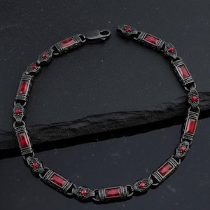 Könnte beinhalten: Ein dunkelgraues Armband mit rechteckigen roten Edelsteinen und dekorativen floralen Akzenten. Das Armband aus Metall hat einen Verschluss. Die Edelsteine sind gleichmäßig über die Länge des Armbands verteilt.