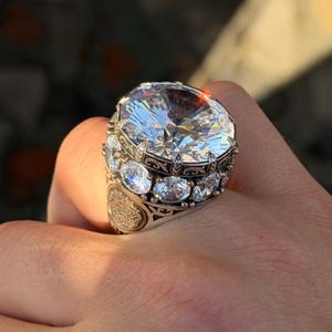 May include: A silver-toned ring featuring a large, round, faceted clear stone at the center, surrounded by a halo of smaller round clear stones. The metal band displays intricate engraved patterns and filigree details along the sides.