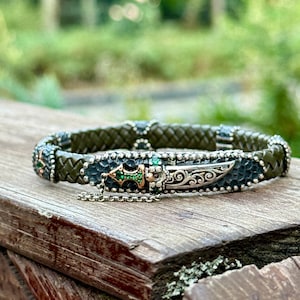 May include: Olive green braided leather bracelet with silver and gold detailing. The bracelet features a decorative dagger design with green and gold gemstones. The bracelet is displayed on a wooden surface.