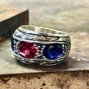 Puede incluir: Un anillo de plata con intrincados detalles grabados presenta una engaste de piedras preciosas rojas y azules. El anillo es una banda ancha con una parte superior redondeada.