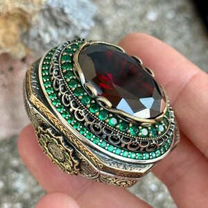 Peut inclure: Une bague ornée avec une grande pierre précieuse ovale rouge foncé. La bague est entourée de petites pierres vertes émeraude rondes et de détails métalliques complexes avec des accents dorés. L'anneau a une esthétique vintage.