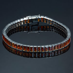 Puede incluir: Una pulsera plateada con dos filas de piedras preciosas rectangulares de color naranja. La pulsera se muestra sobre una superficie oscura y reflectante, resaltando el color de las piedras y el diseño de la pulsera. El cierre es visible.
