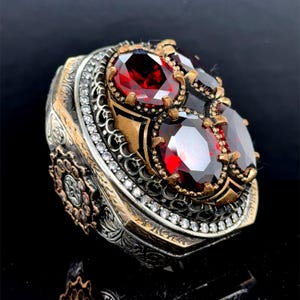 Könnte beinhalten: Ein silberner Ring mit einem detaillierten Design mit vier roten Edelsteinen und einer Reihe von klaren Steinen am Rand.
