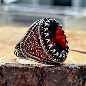 May include: A silver ring with a large, oval-shaped red gemstone surrounded by smaller red gemstones. The ring has intricate details and a textured band.