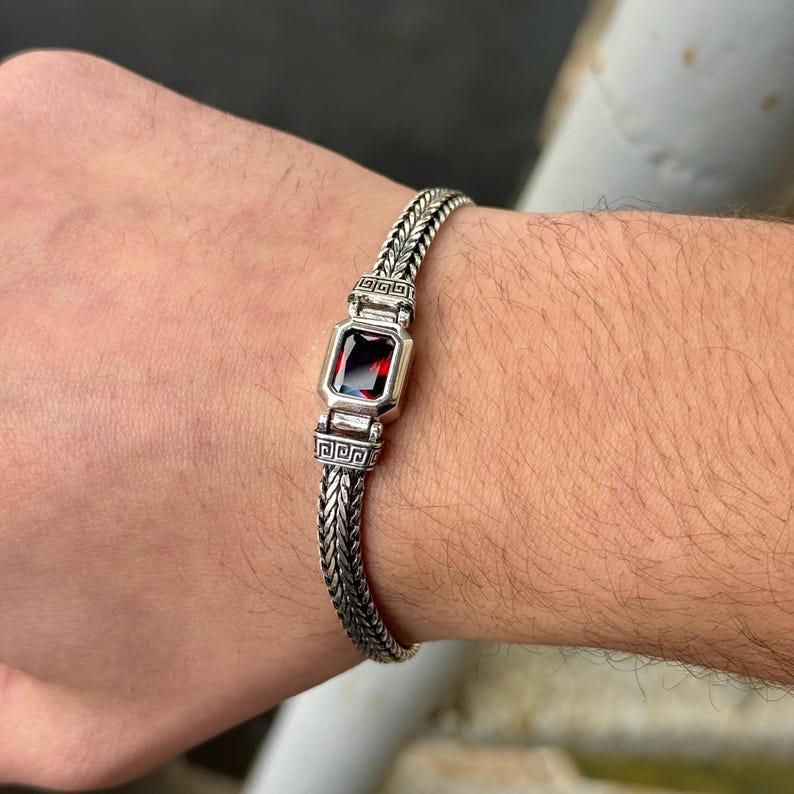 May include: Silver bracelet with a woven chain design. The bracelet features a rectangular, deep red gemstone set in a silver bezel. The bracelet is worn on a wrist.