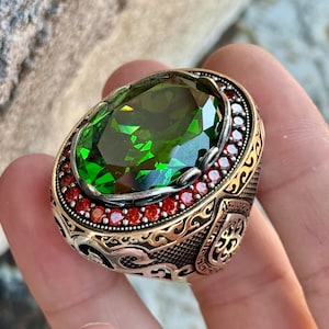 Könnte beinhalten: Ein aufwendiger Ring mit einem großen, leuchtend grünen Edelstein. Der Ring hat ein goldfarbenes Band mit aufwendigem silberfarbenem Filigran und einer Reihe kleiner roter Steine um den grünen Edelstein. Der Ring wird in einer Hand gehalten.
